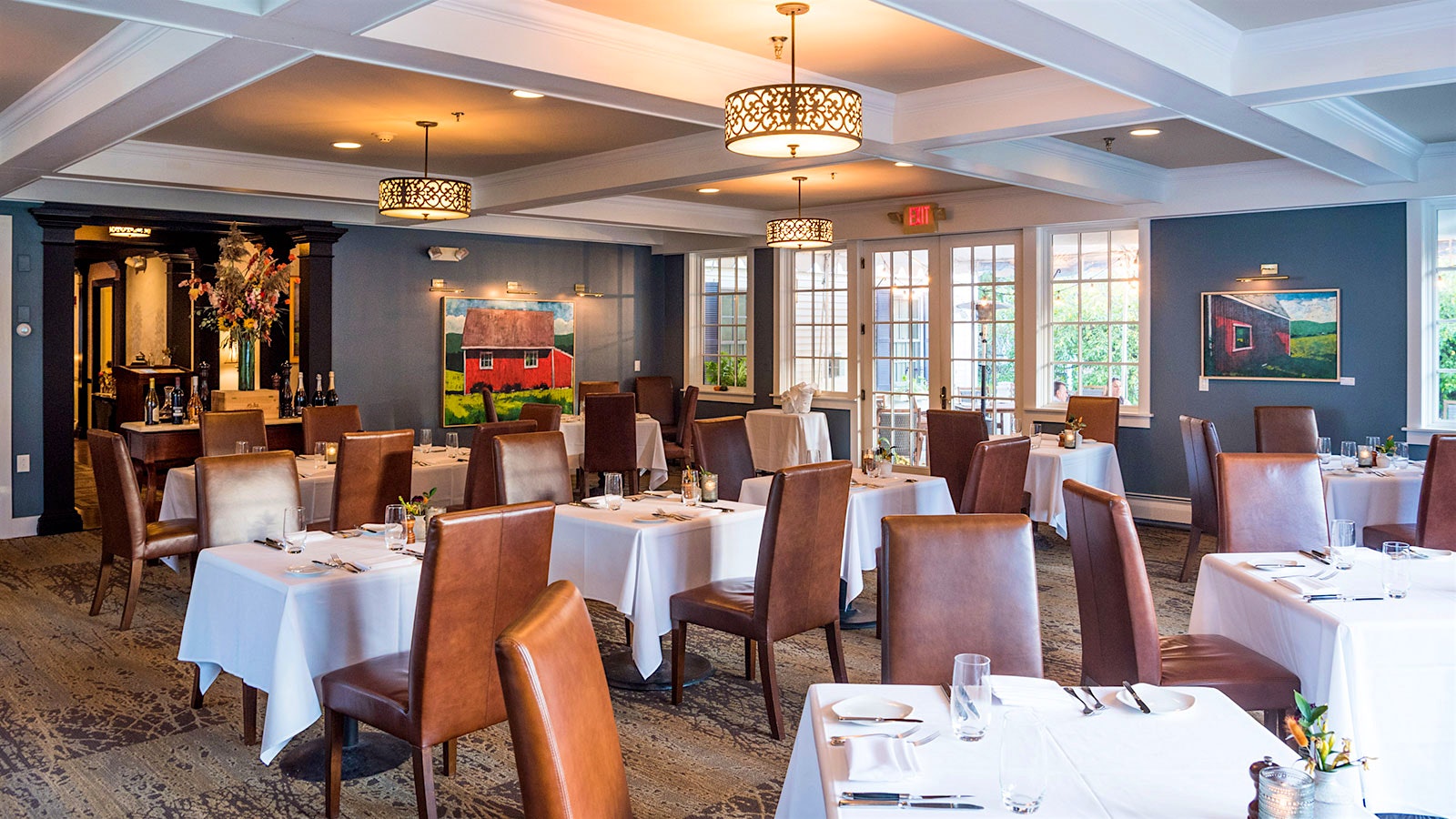 Set tables and leather chairs at the Reluctant Panther, with a painting of a red barn on the wall, a coffered ceiling and round lighting fixtures hanging above; bottles of Champagne and other wine are on a table in the background